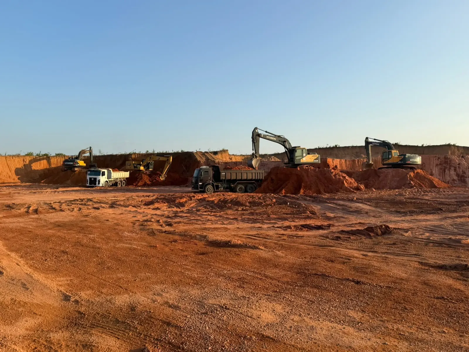 Escavadeiras do Grupo MV Construtora carregando caminhões caçamba em frente de corte de terraplanagem